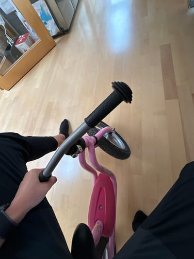 The scene depicts an indoor domestic setting in Grünwald, Germany, observed from a high-angle perspective. The floor is covered with light-toned wooden planks. A pink PUKY brand balance bike is positioned centrally, with its silver handlebars, black rubber grips (one displaying "PUKY"), black seat, silver-rimmed front wheel, and black tire visible. The bike's frame exhibits minor abrasions. A person's hands are gripping the handlebars; the left hand wears a dark wristband. The individual's feet, clad in black socks, are visible on either side of the bike. In the upper left background, a light-colored wooden cabinet or door frame partially reveals a white bag with a blue design and a red "Dermar" box through a glass panel, adjacent to a reflective, metallic surface.