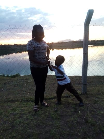 A woman and a young child stand in front of a wire fence near a body of water, likely a lake or pond, reflecting the sky. The woman wears a printed shirt and dark pants, while the child has on a white shirt with colored stripes and dark pants. The child seems to be reaching towards the woman. The background includes a grassy area and distant hills under a cloudy sky, possibly during sunset or sunrise.