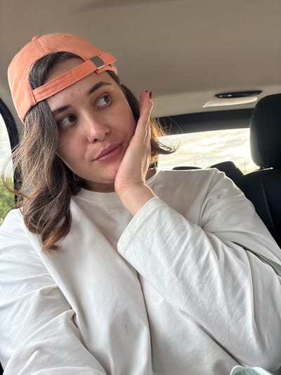 A young woman wearing an orange baseball cap and a white long-sleeved shirt is seated inside a vehicle. She is resting her right hand on her cheek and looking away from the camera. The background shows the interior of a car, with a black headrest visible to the right and a cloudy sky through the window. The lighting suggests it is daytime. There are no identifiable landmarks or indications of the specific location within Zverosovkhoz, Russia.