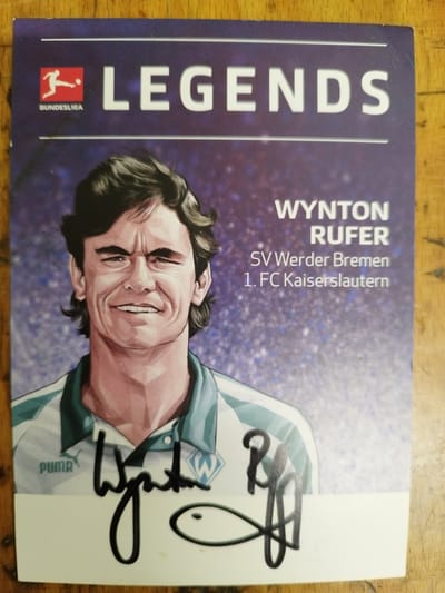 The image is a close-up, top-down view of a collectible card featuring a stylized illustration of footballer Wynton Rufer. The card has a dark purple background with a bokeh effect of white and blue speckles. Across the top, in large white sans-serif font, it reads "LEGENDS". Below this, the name "WYNTΟΝ RUFER" is printed, followed by "SV Werder Bremen" and "1. FC Kaiserslautern" in smaller text. The illustration depicts Rufer from the chest up, wearing a teal and white striped football jersey. His face is rendered with a wide smile and furrowed brow, with dark hair. His signature is prominently displayed in black ink across the bottom of the illustration, above a white border. There is also a small black fish-like doodle next to the signature. The card is placed on a wooden surface. The image does not contain information about the city of Bremerhaven.