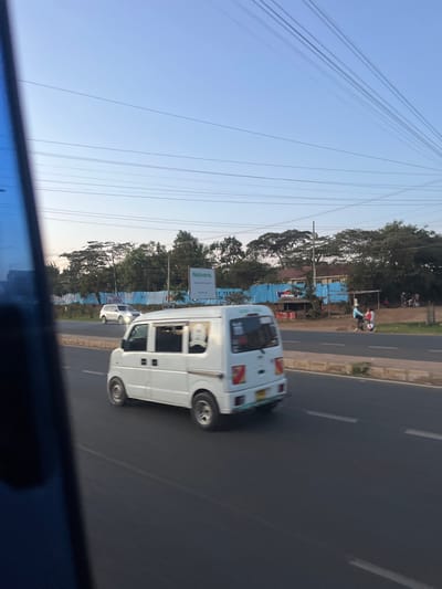 The image depicts a daytime street scene in Biashara ward, Kenya. In the immediate foreground, a blurry blue object, likely a vehicle's interior or window frame, partially obscures the lower left corner.

The central focus is a white, right-hand drive kei van, possibly a mini-bus variant, traveling away from the viewer on a paved asphalt road. The van features black door handles, rear side windows, and a yellow license plate. Red and white reflective markings are present on its rear bodywork. The road exhibits dashed white lane markings.

Across the median, in the opposing lane, a white sedan or hatchback is visible further down the road.

To the right of the main road, on a dirt or unpaved shoulder, two individuals are present. One person, wearing a light blue top, is on a bicycle, while another stands nearby. Further in the background, a small group of people and what appear to be motorcycles are gathered. A small, open-fronted structure with "CROWN" visible on its facade stands along the roadside.

Behind these elements, a continuous line of light blue construction barriers stretches horizontally across the midground. Above these barriers, a large advertising billboard displays "Naivera" prominently and "ST TEXTURE ON" faintly below. Behind the billboard and barriers, dense green trees line the horizon, interspersed with glimpses of a building featuring a reddish roof. The sky is clear and light blue, traversed by multiple horizontal utility lines.
