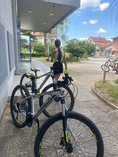 The image depicts an outdoor scene on a paved walkway next to a building. Two bicycles, one in the foreground and one behind it, are parked on the walkway. A young woman with her back to the camera, wearing a grey tank top and black shorts, stands near the bicycles. Her hair is pulled back in a ponytail. In the background, a boy is seen near another bicycle, and further in the distance, houses with red-tiled roofs are visible, characteristic of the village of Ovoshtnik, Bulgaria. The sky is partly cloudy. The environment appears to be a residential area or a small town.