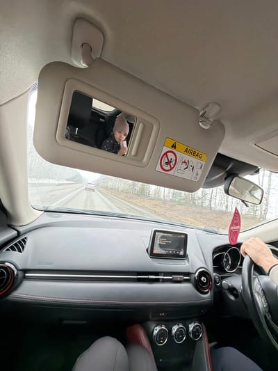 A view from inside a car shows the driver's side. The visor is flipped down and its mirror reflects a baby in a car seat, holding a bottle to their mouth. An air freshener hangs from the rearview mirror. The car's dashboard is visible, including the steering wheel, infotainment screen, and climate control knobs. The location is indeterminable.