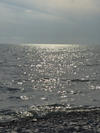 The image depicts a shoreline with a body of water, likely the sea, on the left and a pebbled beach on the right. The water's surface is highly reflective, with numerous points of light scattered across it, indicating the sun is shining brightly and the water is slightly rippled. The sky is overcast and a pale gray, with faint, wispy clouds. The beach is composed of smooth, dark, rounded stones of varying sizes. The perspective suggests the viewer is standing at the edge of the water, looking towards the horizon. There are no people or man-made objects visible in the frame. The scene conveys a sense of natural tranquility and the bright sunlight on the water creates a shimmering, dynamic effect. The time of day appears to be daytime, given the strong sunlight. The location context of Shirokaya Balka, Russia, suggests a coastal environment.