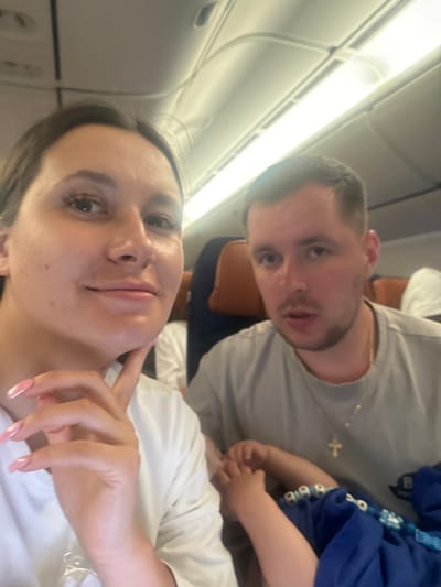 The image captures a selfie-style shot of what appears to be a family inside an airplane cabin. The photograph is oriented vertically, with the subjects appearing rotated 90 degrees clockwise relative to the typical horizontal plane of an aircraft interior.
In the immediate foreground, a woman's face is prominent, taking up the lower half of the rotated frame. She has dark hair, and her eyes are looking directly at the camera. Her expression is neutral to slightly smiling. Her right hand is visible, with manicured nails that are long and have a light pink or white ombré design, resting near her chin. She is wearing a light-colored, possibly white, t-shirt.
Behind and slightly above the woman, a man's face and upper torso are visible. He has short dark hair and is also looking towards the camera, with his mouth slightly ajar as if he might be speaking or just about to. He is wearing a light grey t-shirt and a thin gold chain with a small cross pendant around his neck. His arm and shoulder are visible, suggesting he is seated next to or slightly behind the woman.
To the right of the man, and partially obscuring him, are glimpses of at least one child, possibly two. One child's hands and arms, dressed in a blue top and wearing a blue beaded necklace or accessory, are visible near the man's chest. Another child's head, wearing a white top, is partially seen in the very bottom right corner of the image, close to the woman's side.
The background clearly shows the interior of an airplane. The characteristic curved ceiling with overhead compartments and bright, illuminated light strips running along the length of the cabin are visible. The seats are only partially seen, with brown headrests visible behind the man. The overall lighting is bright, suggesting daytime or well-lit cabin conditions. No text is visible in the image. The scene conveys a casual moment captured during travel, likely a family vacation or trip.