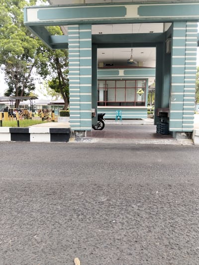 This is an eye-level, medium shot of an outdoor area in Tambontunong, Indonesia. The focal point is a light blue and white striped covered walkway or entranceway, with a dark asphalt road in the foreground. In the background, a building with large windows framed in red is visible, and beyond that, more buildings and lush green trees. There are several objects in the scene: a black motorcycle parked partially within the walkway, a bright blue plastic chair placed in front of the building, and a stack of dark blue water cooler jugs next to one of the walkway's pillars. A yellow sign with black text and a pictogram is affixed to the building's window. The ground inside the walkway appears to be tiled, with a concrete curb painted black and white separating it from the asphalt. The overall scene suggests an institutional or administrative setting, possibly a government building or a school, given the presence of multiple buildings and manicured greenery. The time of day appears to be daytime, with diffused light suggesting it might be overcast or late afternoon. There are no people clearly visible in the frame, implying a quiet or off-hours moment. The environment looks well-maintained. The text on the yellow sign reads "AREA CCTV 24 JAM," which translates to "24-HOUR CCTV AREA" in English, indicating a security measure.