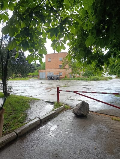 The image depicts an outdoor scene likely in Khorol, Ukraine, on a wet, overcast day. In the foreground, a large, rough-textured boulder sits on a paved surface, positioned to obstruct a red and white striped barrier. The barrier, a typical safety or access control device, is partially lowered and angled. To the left of the boulder and barrier, a strip of green grass abuts a concrete curb, with puddles of water on the pavement indicating recent rainfall.
In the midground, a wide, cracked asphalt area, also wet and reflective, stretches out. A solitary, dark-colored truck with two large wheels and a flatbed trailer is parked in this area, facing towards a large, grey metal garage door. Behind the truck and garage, a multi-story brick building with visible windows stands. Further in the background, more greenery, including trees and bushes, is present. The sky is a muted, cloudy white, suggesting diffused light. A single person, dressed in dark clothing, is visible in the distance near the garage, walking away from the camera. Overhead, the lush green leaves of a tree canopy partially obscure the sky and the upper portions of the buildings. The overall impression is of an industrial or utilitarian area experiencing inclement weather.