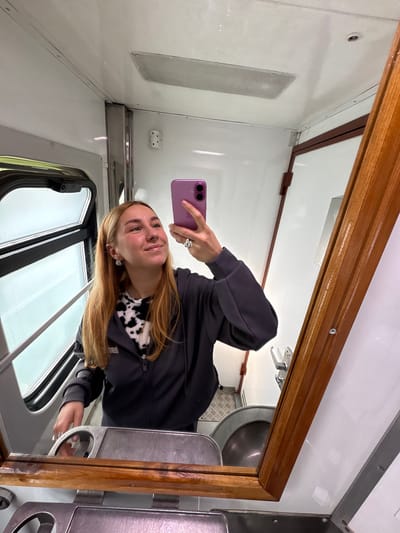 The image shows a woman taking a selfie in a mirror inside what appears to be a train or other passenger vehicle. The walls are white with metal trim. There is a window and a speaker visible. The reflection shows a metal sink. It's difficult to definitively confirm the exact location in Красненскі сельскі Савет, Belarus without additional context or landmarks.