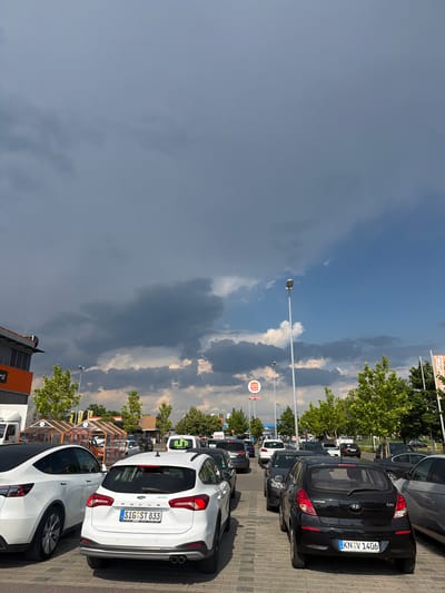 The image depicts a parking lot at a retail or fast-food establishment in Singen (Hohentwiel), Germany, likely near a Burger King, as indicated by a visible sign. The sky is partly cloudy with dark, dramatic storm clouds forming, juxtaposed against patches of blue sky. Several cars are parked in rows on a paved surface. In the foreground, a white Ford Focus station wagon is visible, displaying a German license plate SIG ST 833. To its left is a white Tesla Model Y. Other vehicles, including a black Hyundai i20 with a German license plate KN V 1406, are also present. Trees with lush green foliage line the parking lot, and streetlights are visible. Buildings, including one with an orange facade and a large sign, are situated on the left side of the image. A white van with the word "MUNA" on it is also in the parking lot. The overall scene suggests a commercial area experiencing impending weather changes.