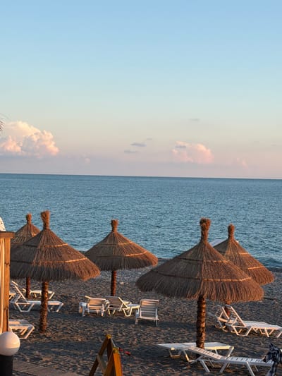 This is a wide shot of a beach in Pitsunda, Abkhazia, at dusk. The sky is a gradient of light blue and pink, with fluffy pink-tinged clouds. The sea is a deep blue, stretching to the horizon. In the foreground, there are several thatched umbrellas providing shade over empty lounge chairs. The sand is dark, and there are some pebbles visible. The scene is calm and serene, with no people present. The lighting suggests the sun is setting or has just set.