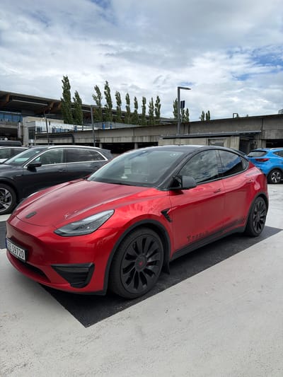 The image depicts a bright red Tesla Model Y parked on a paved surface, possibly a parking lot. The car has a black roof and black accents, including stripes along the lower body and black wheels with red Tesla logos. A license plate with the characters "EE 59720" is visible on the front of the car. In the background, other vehicles are parked, including a dark grey SUV to the left and a blue hatchback further in the distance. Behind the vehicles, there is a multi-level structure with concrete supports and a row of tall, slender trees. The sky above is partly cloudy. The scene is likely at Gardermoen, Norway, given the context of a parking area near a transportation hub like the Oslo Airport. There are no people clearly visible in the image.