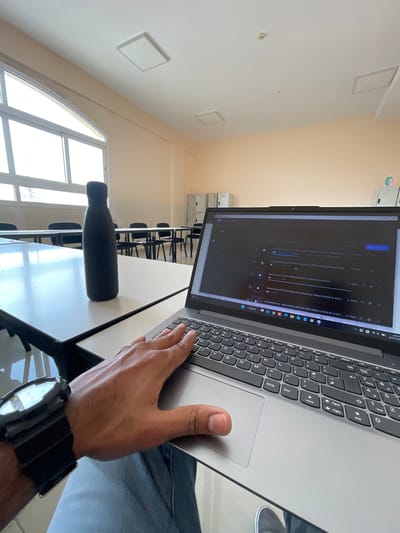 The image depicts a person's hand with a black watch on their wrist resting on a laptop keyboard in what appears to be a classroom or study environment. The laptop screen displays a webpage with a list of video files. In the background, there are several desks and chairs, and a black water bottle is visible on a table. The room has large windows and is illuminated by natural light. The overall setting suggests a modern educational or collaborative workspace. No specific landmarks in Halwan, United Arab Emirates are identifiable in this image.