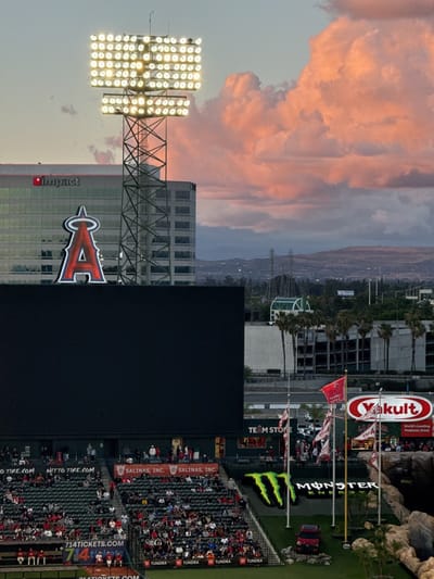 The image depicts a vibrant scene at an outdoor baseball stadium during sunset or dusk in Anaheim, United States. In the foreground, numerous spectators are seated in red stadium seats, facing a large, black scoreboard or video screen. Below the screen, sponsor banners for "NITTO TIRE.COM" and "SALINAS, INC." are visible, along with "714TICKETS.COM" on a railing.

A towering stadium light pole, brightly illuminated with multiple floodlights, stands prominently in the midground. Behind it, a modern building features the "impact" logo and a large, iconic red "A" with a halo, representing the Los Angeles Angels baseball team. To the right, a large green "MONSTER ENERGY" logo is displayed on a sloped grassy area, with a red pickup truck (likely a "TUNDRA") parked nearby. Flags, including one with the Angels logo and another for "Yakult" (which advertises "World's Leading Probiotic Drink"), fly from poles.

The sky above is a notable detail, filled with large, fluffy clouds bathed in warm pink and orange tones from the setting sun, contrasting with the cooler blue sky overhead. In the distant background, a hazy urban landscape with hills and mountains is visible. The stadium lights are fully operational, indicating the transition from day to night. The presence of the Angels logo and "714TICKETS.COM" strongly places the scene at Angel Stadium of Anaheim.