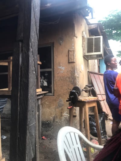 The image shows a scene in Umudike, Nigeria (latitude: 5.481, longitude: 7.546). A person wearing blue clothing and a red shirt stands on a wooden platform, looking off-camera. The platform is supporting a grinding wheel attached to a wooden table. A white plastic chair, a black bag and a piece of wood are visible on the ground, along with other debris. A window, covered by a wooden frame, is visible in the wall behind the wooden platform. The wall is made of tan concrete and has a white, rectangular object attached. The scene is partially obscured by a wooden beam in the foreground, and the top of a tree is visible at the top of the image. The image appears to have been taken from a low angle.