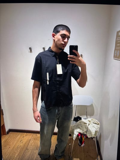 A man in a fitting room takes a selfie. He wears a black short-sleeved button-up shirt with a tag and a small label, along with faded jeans. The fitting room features plain white walls with hooks. A white chair and a clothing rack with folded garments sit in the background. The floor is wooden. A small sign with QR codes is visible on the wall near the ceiling.
