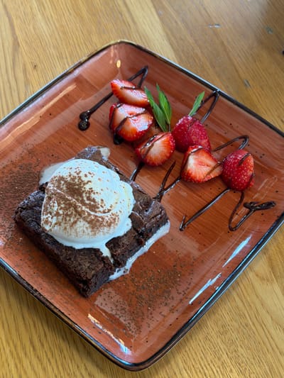A close-up, high-angle shot shows a square, rustic brown ceramic plate on a wooden table. The plate holds a dessert, featuring a dark chocolate brownie topped with a scoop of vanilla ice cream dusted with cocoa powder. Melted white cream oozes from the side of the brownie. The dessert is garnished with several sliced strawberries and a few sprigs of mint, with chocolate sauce drizzled across the plate. The lighting is natural and bright, suggesting an indoor setting, possibly a cafe or restaurant. The wooden table is visible at the top and bottom of the frame. The scene is static, with no people or animals present. The overall impression is one of a beautifully presented dessert.