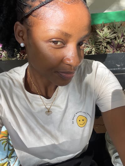 An individual is depicted in a close-up, chest-up portrait shot, positioned outdoors in the Thika Township ward, Kenya. The person, appearing to be female with a dark complexion and dark braided hair, directly faces the camera with a subtle smile. They are wearing a light-colored, short-sleeved t-shirt featuring a yellow smiley face graphic on the left chest area, encircled by the text "DO WHAT MAKES YOU HAPPY". A single white pearl stud earring is visible on the left earlobe. Around the neck, a thin gold-toned chain supports a heart-shaped pendant encrusted with small clear stones. The background, out of focus, reveals green and reddish-purple foliage within a planter or garden bed, with a dark green surface immediately behind the plants. The scene is illuminated by strong, natural sunlight, casting highlights on the subject's skin.