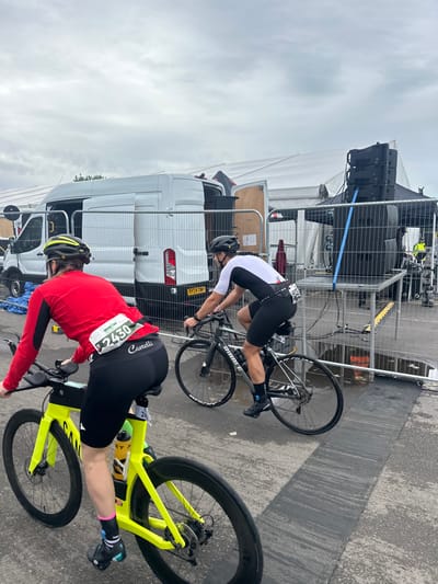 The image depicts an outdoor event scene in Weymouth, United Kingdom, under an overcast sky. Two cyclists are captured from behind in the foreground, appearing to be in motion on a paved surface.
The cyclist on the left wears a red long-sleeved top, black cycling shorts with "Castelli" branding, and a white race bib bearing the "Ironman" logo and number "2430" attached to their lower back. They have a yellow and black helmet, pink and black socks, and black cycling shoes. This individual rides a bright yellow Canyon triathlon bicycle equipped with aero bars and disc brakes, featuring a yellow water bottle in its cage.
The cyclist on the right wears a white and black short-sleeved tri-suit, black cycling shorts, a black helmet, black socks, and black cycling shoes. A race bib with "2000" or similar is partially visible on their lower back. They ride a dark grey Ribble road bicycle with disc brakes.
In the midground and background, event infrastructure is present. A white Ford Transit-style van, with its rear and side doors open, is parked behind temporary metal fencing. Its license plate, "RY24 DWP," is partially visible. Behind the van and fencing, large white canvas marquees are erected. To the right, a multi-tiered black sound system is set up on a temporary platform, secured by blue straps. Puddles are visible on the asphalt ground, particularly near the sound system platform, suggesting damp conditions. A dark rubber mat or ramp is laid across the ground in the midground. Event personnel in high-visibility vests are faintly discernible in the far background near the structures.