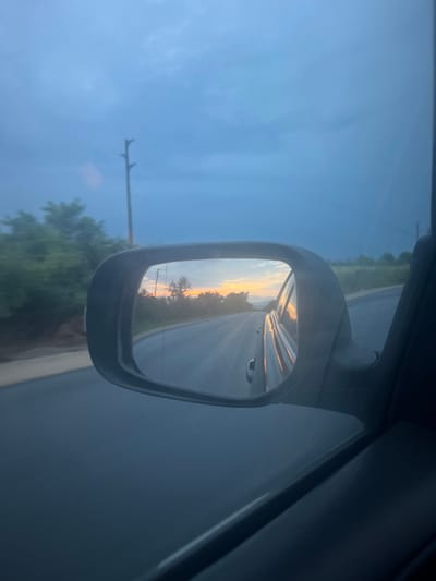 The image captures a view from inside a vehicle, looking out the side window in a portrait orientation. The central focus is the car's side-view mirror, positioned on the right side of the frame.
Within the mirror, a serene landscape is reflected, featuring a road stretching into the distance under the warm, glowing light of either sunrise or sunset. The reflected sky displays a beautiful gradient of soft yellows, oranges, and light blues, indicative of dawn or dusk. Silhouettes of trees line the road in the reflection.
To the left of the mirror, through the car's window, a clear, expansive blue sky dominates, with hints of very light cloud cover. A dark utility pole or power line stands vertically on the far left edge of the frame, with horizontal extensions visible. Below the pole, green trees and roadside foliage are discernible, suggesting a rural or semi-rural setting. The rightmost edge of the image shows a dark portion of the car's interior, likely the door frame.
The image conveys a sense of travel during a tranquil time of day in Buhovtsi, Bulgaria, highlighted by the striking natural light. No visible text is present.