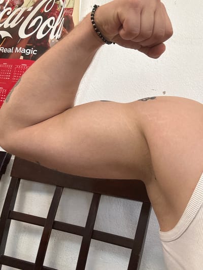 The image shows a close-up of a person's flexed bicep, with a portion of their arm and clenched fist visible. The individual is wearing a beaded bracelet on their left wrist. In the background, a portion of a Coca-Cola advertisement is visible, along with a calendar. A wooden chair is positioned in the foreground, partially obscuring the lower part of the arm. The setting appears to be indoors, with a white, textured wall as the backdrop. There is no discernible indication of the specific location within Oryahovo, Bulgaria, in the provided image.