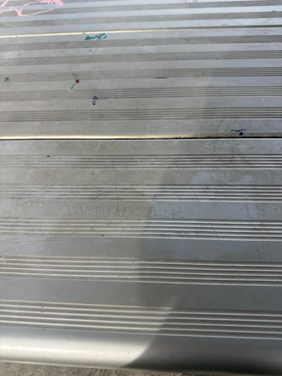The image shows a close-up view of a section of metal bleacher seating. The metal is grey and has a ribbed texture. Etched into the surface are the words "TOMMY H/LR". There are also various other marks, scuffs, and small spots of paint on the metal. The location, Bassendean, Australia, is inferred from the image's origin. No people, actions, or interactions are visible in the frame.