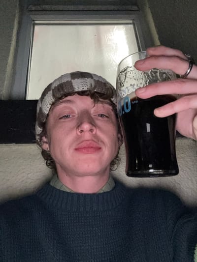 A person wearing a brown and white checkered beanie and dark teal sweater holds a glass of dark beer. The person is fair-skinned with curly hair peeking out from under the hat. They have blue eyes, a nose ring, and a small piercing on their forehead. A window with a pale light is visible behind the person. The person's hand holding the glass has two rings on their fingers.