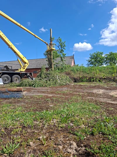 The image depicts a tree trimming operation in Zhabinka, Belarus. A yellow hydraulic crane with a bucket lift is positioned next to a building with a dark roof and red brick exterior. A worker in the bucket is cutting the top branches of a large tree. Cut branches and debris are scattered on the ground near the truck. The sky is blue with scattered white clouds. A dirt path or road runs alongside the tree and building, bordered by vegetation.