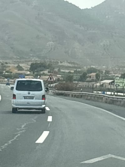 This media file shows a view from a vehicle driving on a road in Lorca, Spain. The main subject is a white van, seen from the rear. The van has a license plate with some visible text, but it is not clear enough to read. The van is traveling on a paved road with white lane markings. In the background, there are hills or mountains with a somewhat sparse, arid landscape. Some buildings and vegetation are visible on the slopes of the hills, suggesting a semi-rural or suburban setting. A triangular road sign is visible on the left side of the road. The sky appears overcast, and the overall lighting suggests it might be daytime, possibly in the morning or late afternoon, with diffused light. The image is taken from a low angle, looking forward and slightly to the left, as if from the passenger seat of a following vehicle. The road curves to the right.