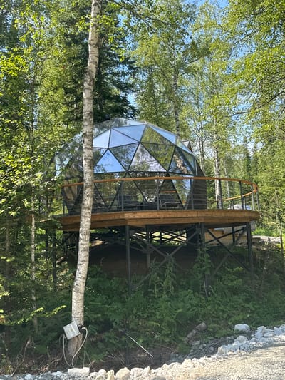 A geodesic dome structure, predominantly made of glass panels, is situated in a wooded area. The dome is elevated on a wooden platform supported by metal stilts. A wooden railing with metal supports encircles the platform. Inside the dome, seating furniture is visible. The surrounding environment consists of numerous trees with green foliage and a clear blue sky. In the foreground, a tree trunk with a metal electrical box attached to it is visible, with wires connected. The ground is covered with gravel and stones. The scene appears to be in the city of Sheregesh, Russia, as indicated.