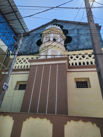 The image displays an upward view of a multi-story building facade in Pedda Kammavaripalli, India, against a clear blue sky. A concrete utility pole occupies the left side of the frame, laden with numerous black electrical and communication wires that crisscross the visual field. A yellow and white paper sign, bearing Telugu script and the number "95152007", is affixed to the pole.
The building's lower section features textured pale yellow walls. A central brown-painted area on the lower facade is vertically striped with three white lines. Two small, rectangular windows with black metal security grilles are observed within the pale yellow sections. Above this, a decorative pale yellow railing with diamond and rectangular openwork patterns is present.
A prominent, ornate tower-like structure, primarily white and pale yellow with arched openings and multiple tiers, extends upwards from the building's central section. The uppermost visible part of the building consists of a textured, weathered blue-green wall beneath a dark, possibly corrugated, roof edge. To the far left, a corrugated metal roof covered with blue material from an adjacent structure is partially visible. No individuals or specific actions are discernible within the frame.