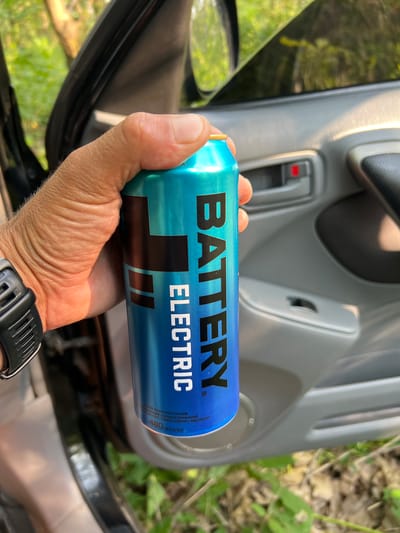 In the city of Novohnide, Ukraine, a person's hand is holding a blue and teal can of "BATTERY ELECTRIC" energy drink. The hand, showing wrinkles and some arm hair, is wrapped around the can, obscuring part of its label. A black watch with a digital display is visible on the person's wrist. The background suggests the interior of a car, with a grey door panel, a red door lock button, and a black armrest. Sunlight casts highlights on the car's interior surfaces. Outside the car, blurred green foliage and dry ground are visible, indicating an outdoor setting. The can itself has black lettering and a black logo with abstract shapes. Text in a smaller font, partially visible at the bottom of the can, indicates "480 ml [ml]". The overall scene depicts a moment of refreshment taken in a vehicle, possibly during an outdoor excursion.