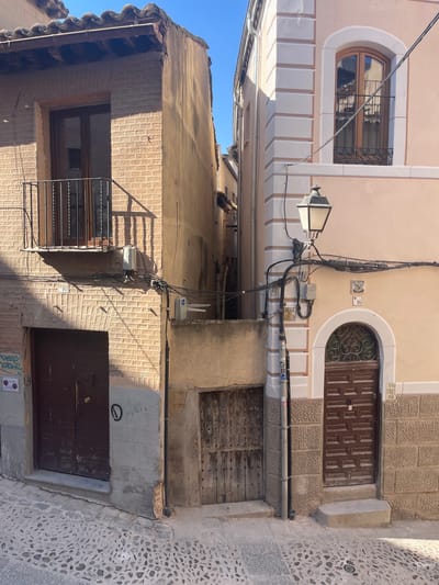 The image depicts a narrow cobblestone street in the city of Toledo, Spain, with two buildings meeting at a corner. The building on the left is constructed of light-colored brick and features a dark wooden door at street level and a balcony with a black metal railing above. The building on the right is painted a pale peach color and has decorative light beige trim around a large arched window with a wooden frame. A dark wooden door with decorative metalwork above it is visible on this building. A traditional street lamp with a black metal post and decorative scrolled arms is positioned between the two buildings. Various electrical conduits and junction boxes are attached to the facades of both structures. The sky above is clear and blue.