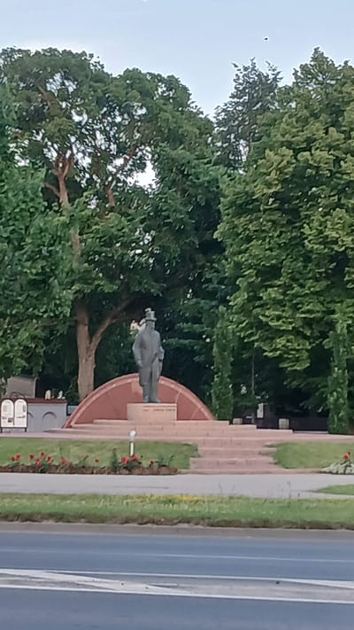 The image shows a full-body statue of a man standing outdoors, possibly in a park or public square. The statue is dark in color and appears to be made of bronze or a similar material. The man is wearing a suit and a hat, and is holding something in his hands, possibly flowers. He is standing on a raised platform or monument, which is made of reddish-brown stone. The monument has a circular or semi-circular base, with tiered steps leading up to it.
Behind the statue, there are large trees with green foliage, suggesting a warm season. The sky is visible above the trees, and it appears to be overcast or hazy. To the right of the monument and trees, there is a paved road with white lane markings, indicating it is a street or highway. A grassy area with some small red flowers is visible between the monument and the road.
The image is oriented vertically, with the top of the scene on the left and the bottom on the right. This orientation might be due to how the photo was taken or how it is being displayed.
There are no discernible emotions conveyed by the statue, as it is a static representation. The time of day appears to be daytime, judging by the lighting and the visibility of the sky. The weather seems to be mild, with the presence of lush greenery and flowers. The location is identified as Jelgava, Latvia.
There is some text visible on a plaque on the monument, but it is too blurry to be read.