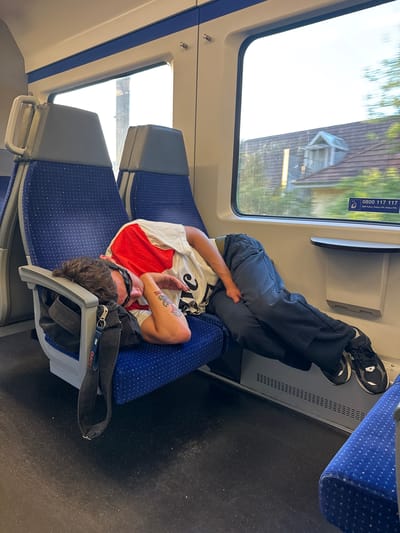 A man is sleeping on a train, slumped over in his seat. He is wearing a red and white t-shirt and dark pants. His head is resting on the armrest, and he has sunglasses on. A black bag is hanging from the armrest. The train seats are blue with small white dots. Through the window, houses with tiled roofs and trees can be seen. A sticker on the window shows a phone number, "0800 117 117", and "SBB Police, Police CFF, Police FFS". The floor of the train is dark and appears to be made of linoleum. This scene likely takes place on a SBB (Swiss Federal Railways) train. Given the presence of the SBB phone number and the scenery, it is plausible that this train is traveling through or near Biel/Bienne, Switzerland, as Biel/Bienne is a major railway hub in Switzerland and a bilingual city where German and French are spoken, hence the "CFF" (Chemins de fer fédéraux suisses) and "FFS" (Bundesbahnen) designations on the sticker.