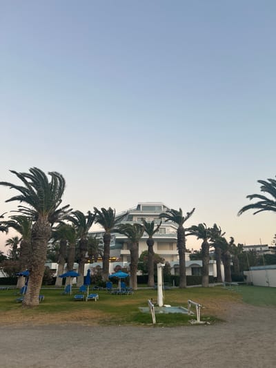 The image depicts a resort scene in Rhodes, Greece. A modern, multi-story hotel with white balconies and a distinctive angular roof is visible in the background. Palm trees are scattered throughout the property, with some in the foreground casting shadows on a well-maintained lawn. Several blue lounge chairs and umbrellas are set up on the grass, suggesting a recreational area. A gravel path runs in front of the lawn. The sky is a clear, pale blue, indicating either early morning or late afternoon. No people are visible in the image.