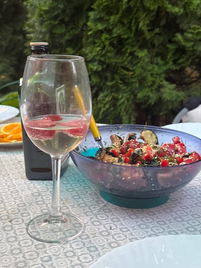A glass of white wine with strawberry slices sits on a table covered with a patterned tablecloth. To the right of the glass is a blue bowl filled with a salad of roasted vegetables, including eggplant, tomatoes, and peppers, garnished with sesame seeds and dill. Behind the wine glass, a dark bottle, possibly olive oil or wine, is visible. In the background, lush green foliage suggests an outdoor setting, possibly a garden or patio. A white plate with orange slices is partially visible to the left of the wine glass. The scene is likely located in Pribrezhnyi, Russia, given the context.