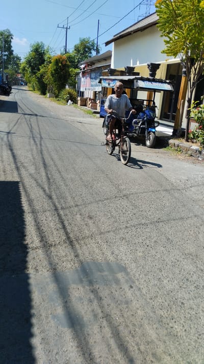 The image captures a street scene in Kramat Jegu, Indonesia, on a bright, sunny day. In the foreground, an older man with a bald head and wearing a striped short-sleeved shirt is riding a red bicycle. The bicycle appears to be a child's or a small adult bicycle, with white tires and chrome handlebars. The man is looking forward, and his feet are positioned on the pedals. To the right of the bicycle, a blue motorcycle is parked. The street is paved with asphalt and shows signs of wear. Numerous tree shadows are cast across the road from overhead power lines and unseen trees. In the background, there are buildings, including a white building with a tiled roof and a smaller structure with a corrugated metal roof. Several trees with green foliage are visible along the side of the road, and some orange and red flowers are growing near the white building. To the left of the street, parked motorcycles are visible. Signs are posted on the buildings in the background, with some text visible but not clearly legible. The sky is a clear blue.