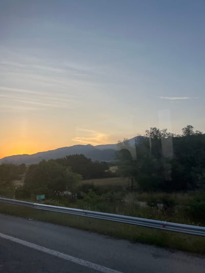 The image displays a panoramic landscape captured from a moving vehicle at either sunrise or sunset, with the location identified as Amaxades Municipal Unit, Greece. The sky occupies the upper two-thirds of the frame, transitioning from a pale blue at the top to a vivid orange-yellow at the horizon, where the sun's light is intensely concentrated on the left side. Thin, horizontal stratiform clouds are present in the upper sky, and a more distinct, elongated cloud appears in the mid-right. A faint, linear white contrail is visible in the upper left quadrant of the orange sky.

Multiple layers of mountains are silhouetted against the bright horizon, appearing as dark blue-grey masses with an undulating profile. Below the mountains, the midground consists of dense, dark green deciduous trees interspersed with lighter-toned fields or patches of dry vegetation. The image shows noticeable reflections, appearing as vertical streaks and faint, semi-transparent overlays across the landscape, particularly obscuring parts of the trees in the center-right.

The foreground features a segment of a paved road, likely asphalt, with a solid white line marking its left edge. A metallic, silver-grey guardrail extends horizontally across the lower portion of the frame, separating the road from a grassy embankment. Behind the guardrail and partially concealed by foliage, a green rectangular road sign with white lettering displays "H.P. 534". A small, indistinct red object is visible on top of the guardrail near the center-right. No individuals are present in the scene, and no specific actions or interactions are depicted.