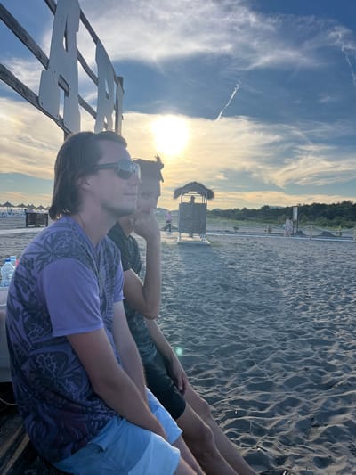The image shows two men sitting on a wooden bench on a sandy beach. The man in the foreground is wearing sunglasses, a purple t-shirt with a black pattern, and light blue shorts. The man behind him is wearing a dark t-shirt and dark shorts. They are looking out towards the sea. In the background, there are beach umbrellas, a lifeguard tower, and vegetation. The sun is setting, casting a warm glow on the scene. The sky is partly cloudy with a contrail visible. The location is identified as the city of Pistula, Montenegro.
