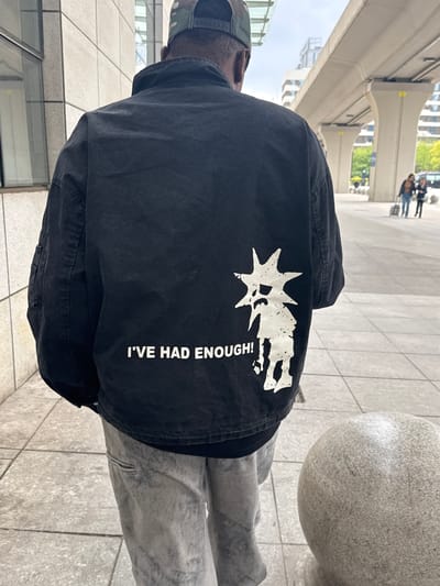 The image shows a person wearing a black jacket with a white graphic print on the back. The print consists of a stylized sun or starburst shape, a figure that appears to be holding their hands up, and the text "I'VE HAD ENOUGH!". The person is also wearing a camo-patterned cap and gray pants. They are standing on a paved area with large tiles and there are buildings and elevated structures in the background. The scene appears to be in an urban environment, possibly in Shenhe District, China, as requested.