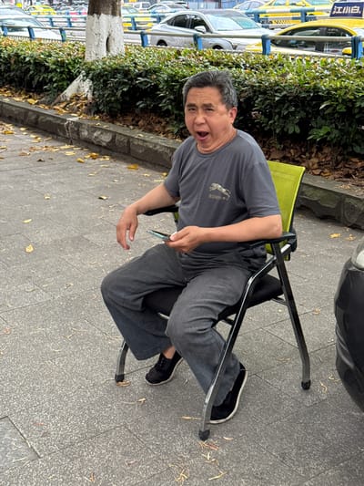 A man sits in a folding chair on a sidewalk in what appears to be 龙溪街道, China. He wears a gray t-shirt and gray pants, with black shoes. He is holding a cell phone in his left hand and gesturing with his right. Taxis are parked in the background, partially obscured by bushes. The sidewalk is gray and shows some fallen leaves. A car is partially visible in the upper right of the frame.