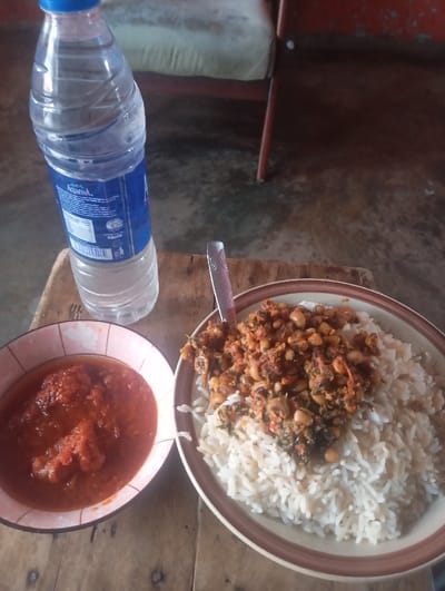 The scene depicts a meal set on a wooden surface in an indoor location within Okere-Urhobo, Nigeria. A partially filled Aquafina plastic water bottle with a blue cap is positioned vertically on the left. To its right, a large, round plate contains a portion of white rice topped with a stew-like mixture of black-eyed peas, leafy green vegetables, and a red sauce. A silver-colored spoon is inserted into the stew. To the left of this plate, a smaller bowl with a pink floral rim holds a reddish-orange stew, containing visible pieces of protein. The background is out of focus, revealing a dark concrete floor, a wooden chair with a light-colored upholstered seat, and a reddish-orange wall. No individuals are present in the image.