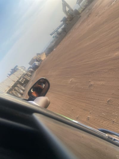The image captures a view from inside a vehicle, looking out towards an expansive, unpaved area. The original image is rotated 90 degrees clockwise; when viewed in a corrected orientation, the perspective is from the driver's or passenger's side of a vehicle, looking out the right-hand window.
In the immediate foreground, to the bottom right, parts of the vehicle's interior are visible, including a dark-colored dashboard or door panel. A side-view mirror protrudes from the vehicle's exterior. Reflected in this mirror is a portion of a person's arm or hand, with a distinct orange or reddish-brown sleeve, suggesting someone is inside the vehicle.
Beyond the side mirror, slightly further back and extending towards the center-right of the frame, is a large multi-wheeled vehicle, possibly a truck or a piece of heavy equipment like a trailer, which the viewing vehicle appears to be alongside or passing.
The dominant feature of the scene is a vast, open expanse of light brown, sandy, or dusty ground, which stretches from the mid-ground into the distant background. Numerous parallel tire tracks and other marks are visible on this unpaved surface, indicating regular vehicle movement. The ground appears dry and uneven in places.
In the far background, along the left side and towards the top of the frame, a significant amount of industrial or construction equipment is visible. This includes structures resembling cranes, excavators, and other large machinery, suggesting an active work site such as a quarry, mining operation, or a major construction project. The sky above this industrial landscape is bright and appears somewhat hazy, indicating either a sunny day with dust in the air or atmospheric haze.
The overall impression is that of a vehicle traveling on a dirt road through an active industrial or heavy-duty operational area during daylight hours. No discernible text is visible in the image.
