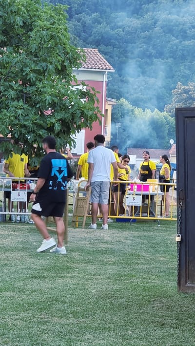 The image shows a group of people gathered outdoors on a grassy area. In the background, there is a dense forest and a building with a red tiled roof. Smoke is visible in the air, suggesting some sort of activity like cooking or a small fire.

Several people are wearing yellow t-shirts, some with text that appears to be "BLATISM". One person in the foreground is wearing a black t-shirt with a blue graphic that resembles the Off-White logo and white shorts. Another person is wearing a white t-shirt and grey shorts. The overall scene suggests an outdoor event or gathering, possibly a festival or celebration given the festive atmosphere and the yellow t-shirts which might be uniforms.

The time of day appears to be daytime, with natural light illuminating the scene. The weather seems pleasant, as people are outdoors in casual attire. The location context provided is Sant Joan Les Fonts, Spain. There is also a yellow fence-like structure with some people standing behind it, possibly at a stall or serving area. A lock is visible on a chain attached to this structure. A green tree with large leaves is prominent on the left side of the image.