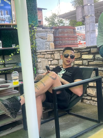 The image depicts an outdoor daytime scene in Касапите, Bulgaria, centered on a young adult male seated in a black metal-framed chair. The male, with short dark hair and wearing sunglasses, a black t-shirt with a partially visible white graphic, and black shorts, has script and pictorial tattoos on both legs and arms; a script tattoo reading "I need god" is on his right thigh, and a feather-like design is on his left shin. He holds a black smartphone in his right hand, facing slightly towards the viewer with his mouth open. His left leg is crossed over his right. Black sneakers, showing some dirt, are on his feet, and a green and black ankle device is visible on his left ankle.

To his left, a table constructed from light-colored wooden pallets holds a clear plastic cup containing a yellow liquid, a brown wallet, and a black smartphone. Behind the table, a dark shelving unit adorned with artificial green ivy displays a small white lantern, a small glass, and a blue box. A vintage-style television, painted white and brown, sits on the top shelf, displaying an advertisement with two male soccer players (numbers 32 and 17 visible) and the "inbet" logo, along with Cyrillic text "HOBO" and "HAC".

The immediate background features a rustic stone wall constructed from irregular light brown and gray stones. Above the wall, a large weathered wooden barrel secured by metal bands is prominent. Further back, a metal utility pole with an attached electrical box and a partially visible advertisement poster for "АВТО НА СА" (featuring a red car) are present. Green foliage and trees are visible in the distance under a bright sky, indicating daylight. A person's upper torso and arm in a light green shirt are partially visible on the far right edge of the frame. A cigarette butt and a small coin lie on the light-colored paved ground near the subject's chair.