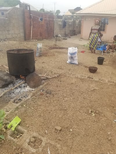 The image shows a dirt yard located in the city of Pati Ridge, Nigeria, at the coordinates (latitude: 7.81, longitude: 6.704). In the background, there is a concrete wall with a rusted metal gate, and a building with a tin roof. Clothes are hanging on a line strung between the two. In the center of the image, a woman wearing a checkered long-sleeved dress stands with her back to the camera. She is near a woman and a girl sitting on a blue chair. There are two large white bags of fertilizer lying on the ground in front of the woman. A large black metal barrel with a small fire burning underneath it is in the foreground. There is a small plastic bucket on the ground next to the barrel. Two more buckets, one dark gray and the other blue, can be seen near the woman in the center.  The ground is covered in dirt and there are a few scattered rocks and sticks. The vegetation in the image is sparse, but a few small plants are growing near the edge of the yard.  The overall scene is quiet and peaceful.