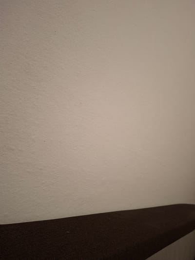 This is a close-up, indoor shot of a textured beige wall above a dark brown upholstered surface. The wall occupies the majority of the frame, showing a slightly uneven, porous texture with a few subtle specks of darker color. The brown surface, which appears to be a piece of furniture like a sofa or bench, is in the lower portion of the image, curving slightly upwards from left to right. The lighting creates a soft gradient on the wall, with the top appearing brighter than the bottom. There are no people or animals visible in the image. The scene is domestic or public interior, likely a waiting area or lounge. It is impossible to determine a specific location in Ilava, Slovakia, from this image alone.