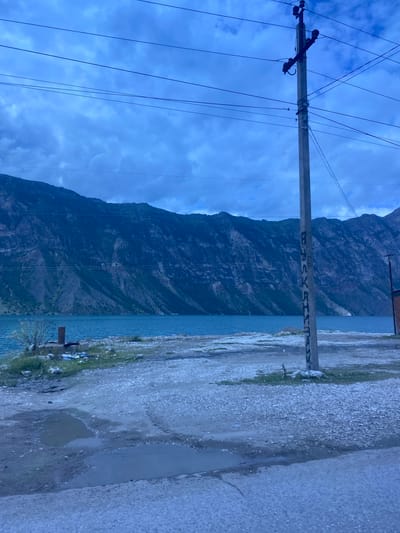 The image depicts a dusky, overcast day in the vicinity of the Irganay reservoir in Irganay, Russia. A concrete utility pole with electrical insulators and wires is prominently featured in the foreground, with Cyrillic writing on its surface. The pole stands on a dirt and gravel embankment that slopes down to the bright blue waters of the reservoir. Beyond the reservoir, a massive, dark, and rugged mountain range dominates the background, its steep slopes etched with shadows and some sparse vegetation. The ground in the immediate foreground is littered with debris, including plastic bags and other refuse, and shows evidence of puddles and tire tracks. A small, unkempt patch of grass is visible near the base of the utility pole, with a low concrete structure and more scattered trash. To the far right, a portion of a brick building is visible. The overall impression is one of a somewhat neglected, natural landscape adjacent to man-made infrastructure.