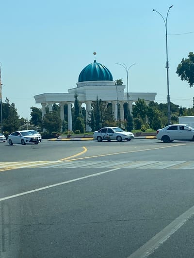 In Andijan City, Uzbekistan, a wide street with a pedestrian crossing is visible in the foreground. Cars are driving on the road, with a white sedan in the left lane of the pedestrian crossing and another white sedan further down the street. In the background, a large white building with a teal dome topped by a gold star stands prominently. The building is supported by numerous white columns. Lush green trees surround the structure, and two streetlights are present on the right side of the image. The sky is clear and blue.