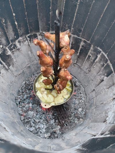 A circular, brick-lined pit, likely a tandoor or similar oven, contains glowing embers at the bottom. Suspended above the embers is a metal skewer holding several pieces of browned chicken. Directly beneath the chicken, a shallow pot is filled with cut potatoes and liquid. The surrounding area is composed of rough, gray stone. The scene appears to be outdoors, with no other structures or people visible. The image was captured in Востец Снт, Russia.